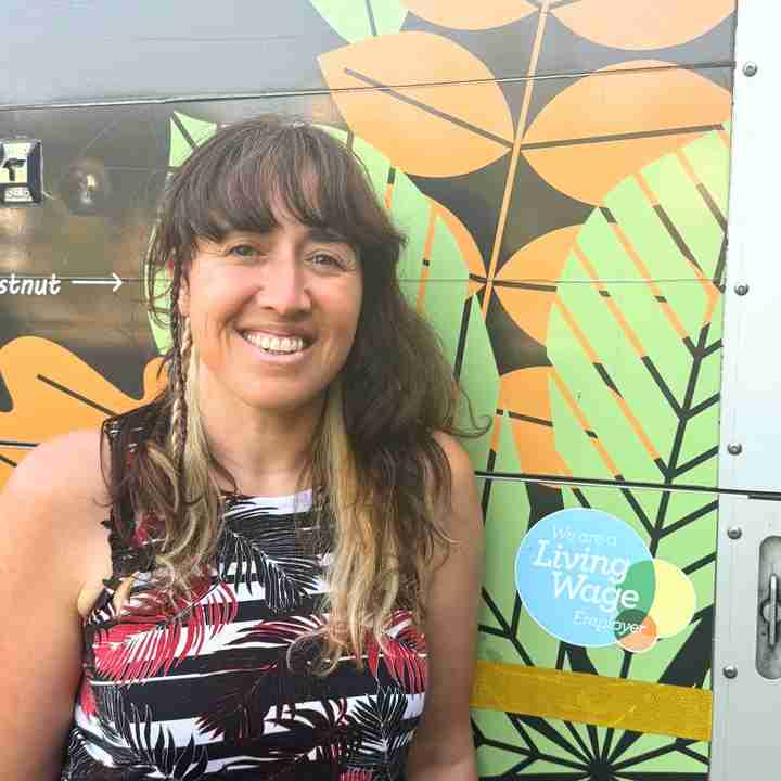 Smiling woman with long brown hair stands in front of a colourful leaf-patterned wall with a "Living Wage Employer" sticker visible.