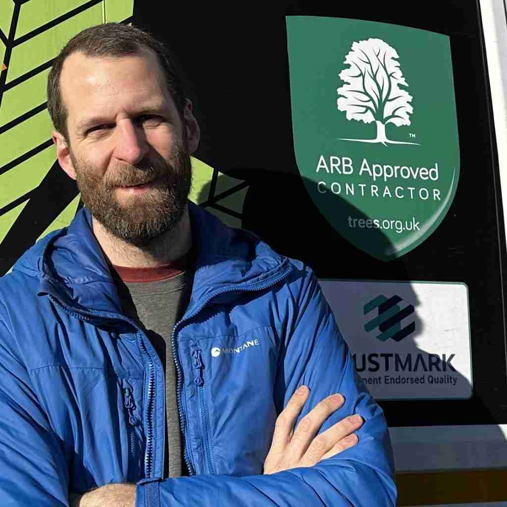 Man in blue jacket standing in front of a sign for an ARB Approved Contractor and TrustMark endorsement, with a green tree logo visible.