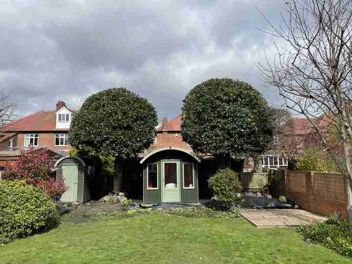 A garden with a small round building in the centre, flanked by two large trimmed trees, with houses and a cloudy sky in the background.