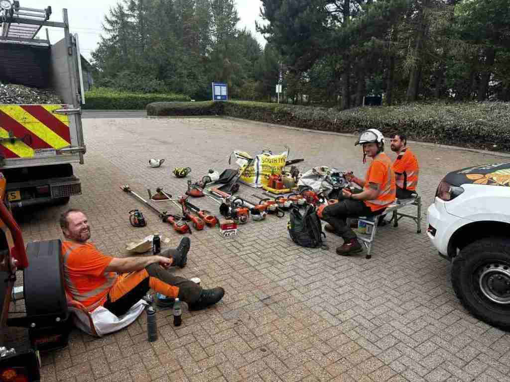 Three workers in orange safety clothing sit on a paved area with landscaping tools and equipment laid out around them, taking a break near parked vehicles.
