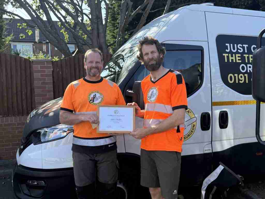 Two men in orange shirts stand in front of a van, holding a tree surgery certificate together and smiling at the camera.